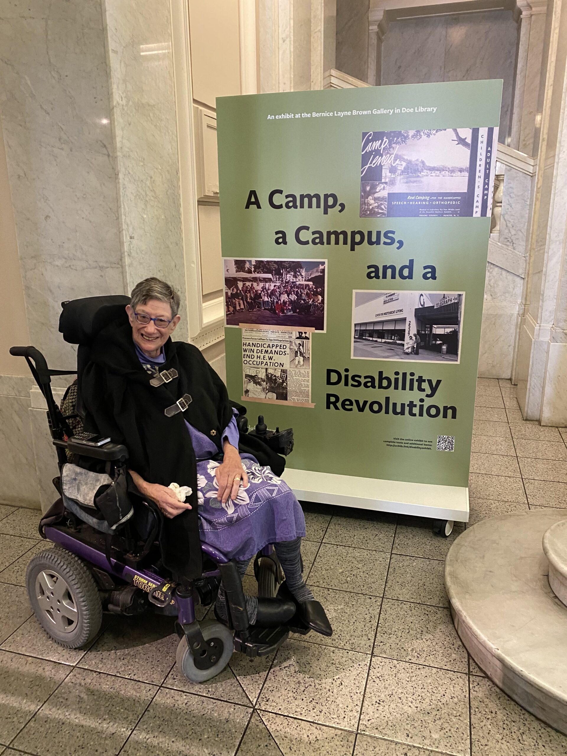 Denise Sherer Jacobson smiling next to a banner that says "A Camp, a Campus, and a Disability Revolution"